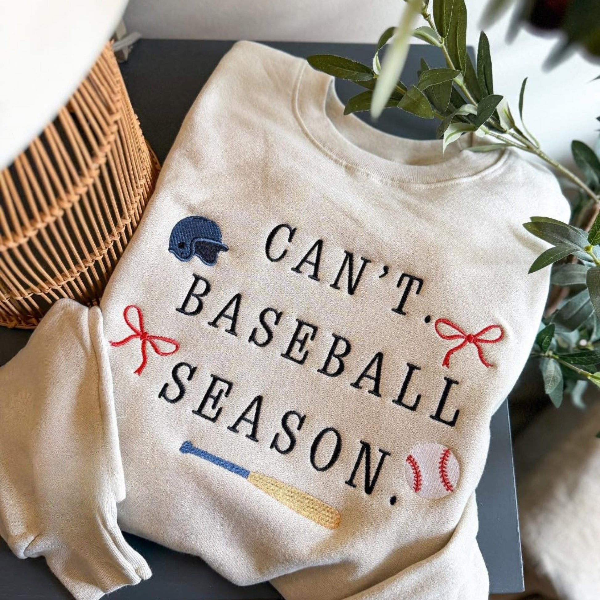 White sweatshirt with 'Can't. Baseball Season.' text and sports-themed graphics on a surface with a plant in the background.