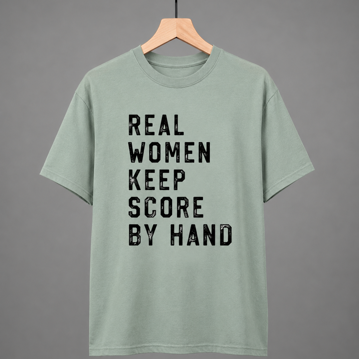 Green t-shirt with 'Real women keep score by hand' text on a hanger against a gray background