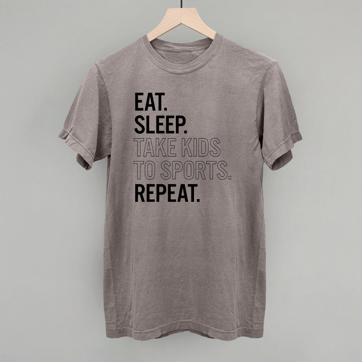 Gray comfort colors  t-shirt with text 'EAT. SLEEP. TAKE KIDS TO SPORTS. REPEAT.' on a hanger against a gray background