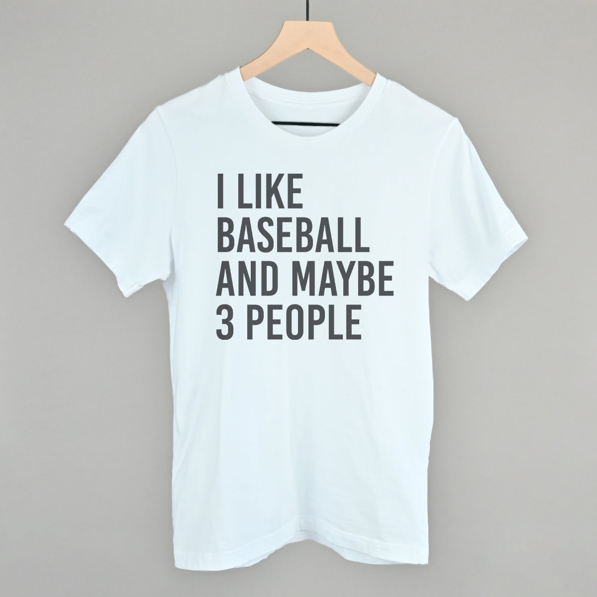 White t-shirt with 'I LIKE BASEBALL AND MAYBE 3 PEOPLE' text on a hanger against a gray background