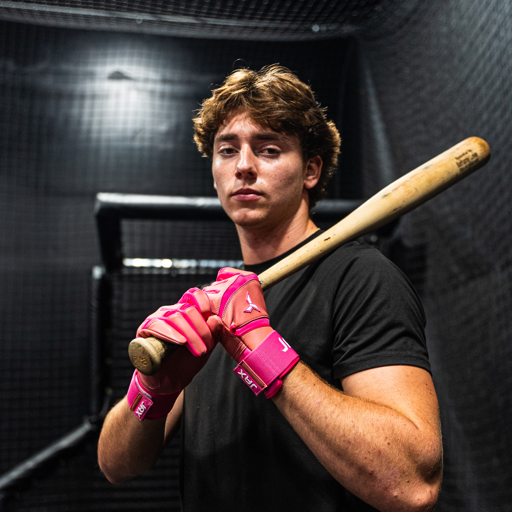 Person holding a baseball bat with pink batting gloves in a dark indoor setting