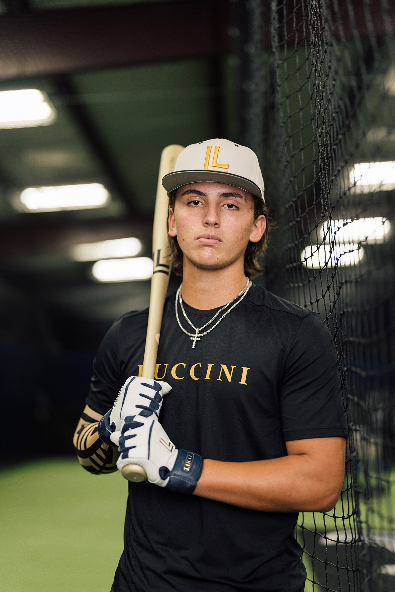 Person holding a baseball bat in front of a batting cage, wearing a black shirt with 'Lucchini' branding.