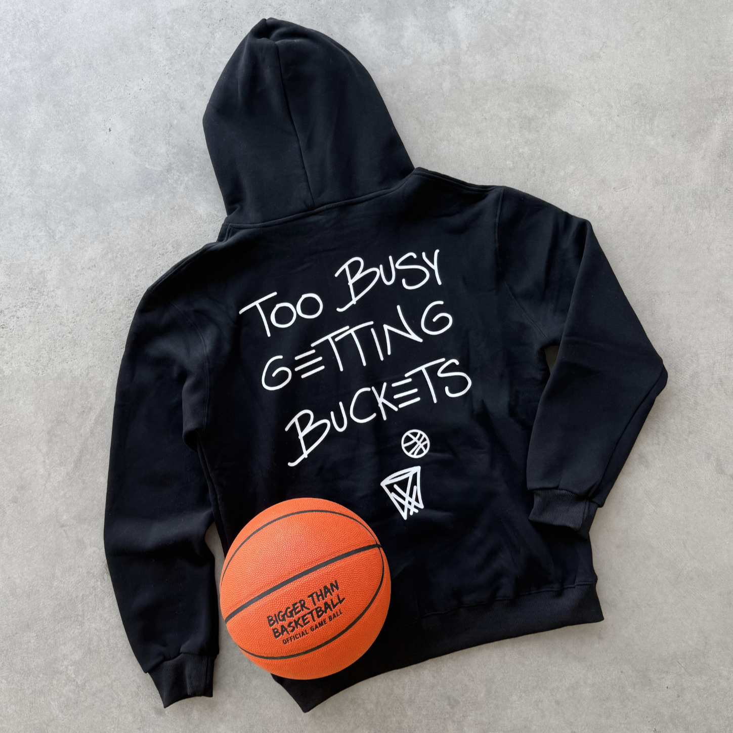 Black hoodie with 'Too Busy Getting Buckets' text and a basketball on a gray surface