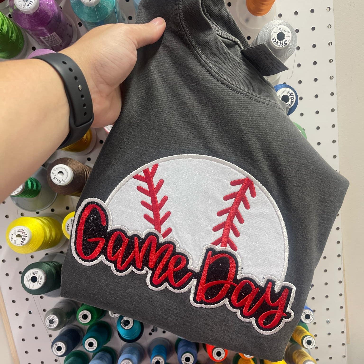 Gray sweatshirt with 'Game Day' patch held in front of a pegboard with spools of thread.