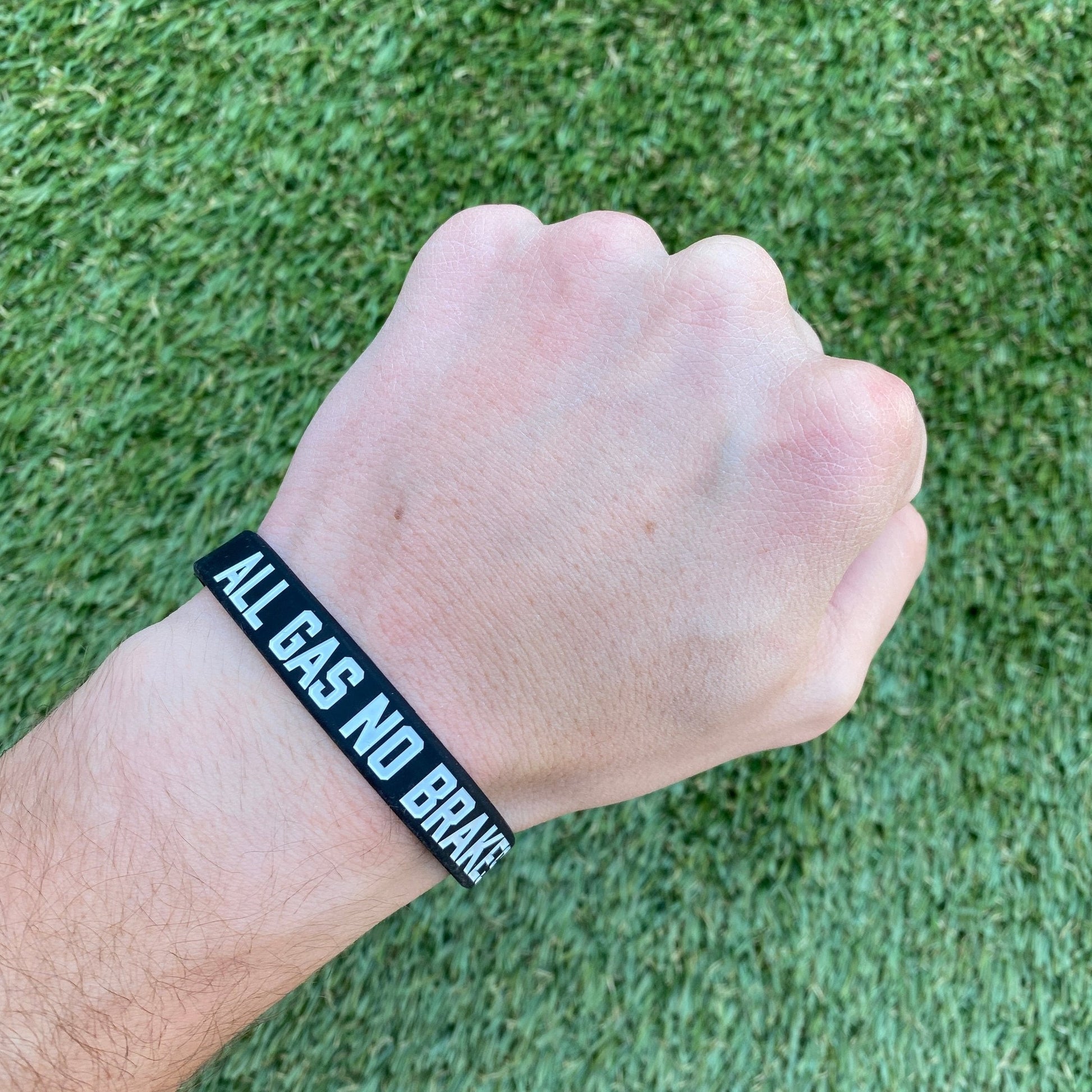 Hand wearing a black wristband with 'ALL GAS NO BRAKE' text on a grass background