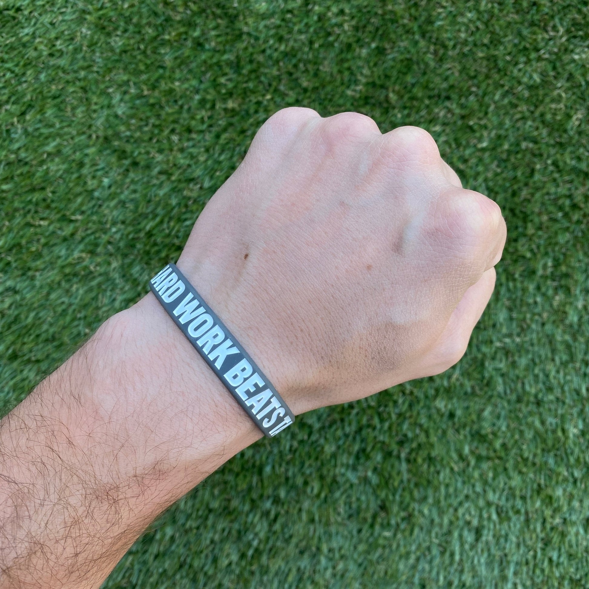 Hand wearing a wristband with 'Hard Work Beats TALENT' text on a grass background