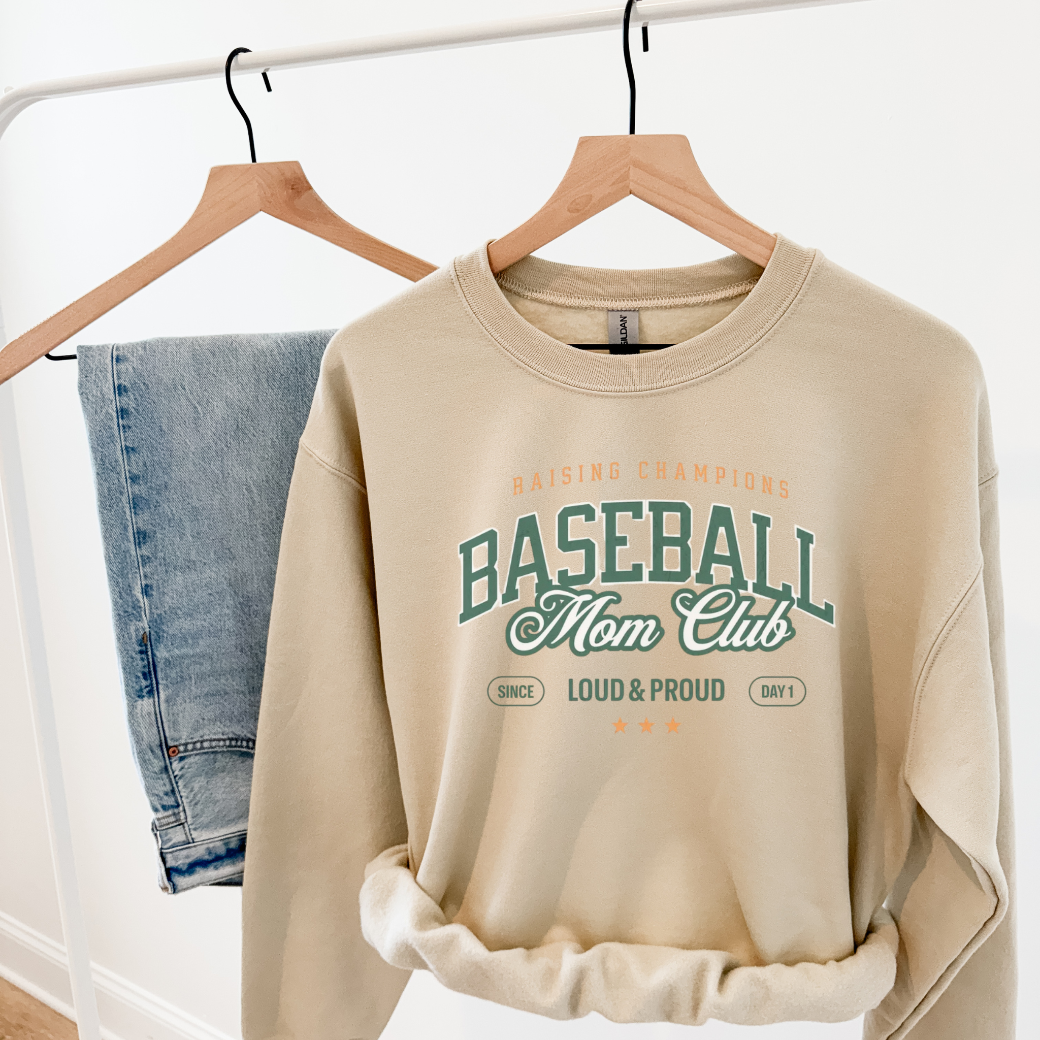 Collection of Beige sweatshirt with 'Baseball Mom Club' text on a hanger with jeans in the background. in a gallery layout